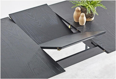 A dark black wood-look extendable dining table shown partially unfolded, with a small potted green plant and two beige decorative objects on top.