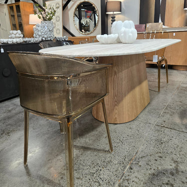 A modern dining table with a white marble top and a sculptural light wood pedestal base, paired with a metallic bronze chair in a furniture showroom.