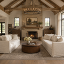 A rustic, high-ceilinged living room with exposed wood beams, a stone fireplace, and a round wood coffee table centered between two off-white slipcovered sofas on a large patterned rug.