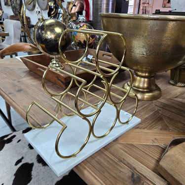 A gold metal wine bottle rack with a spherical accent on top, displayed on a white rectangular base on a wooden table in a home decor store.