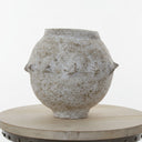 An antique-style textured ceramic pot with a distressed, weathered finish, displayed on a round wooden pedestal against a white paneled wall.