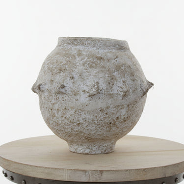 An antique-style textured ceramic pot with a distressed, weathered finish, displayed on a round wooden pedestal against a white paneled wall.