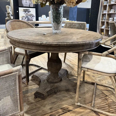 A round rustic wooden dining table with a pedestal base, surrounded by woven and light wood chairs, decorated with a large dried floral arrangement in a glass vase.