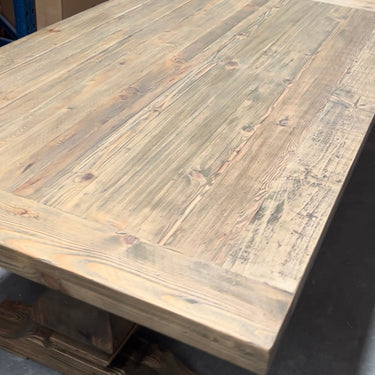 A rustic rectangular wooden dining table with a thick plank top and trestle-style base, photographed in a warehouse or storage area with cardboard boxes in the background.
