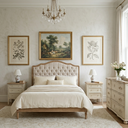 A classic bedroom scene featuring a light wood and tufted bed dressed in cream bedding, flanked by matching nightstands with lamps. Above the bed, three framed botanical and landscape prints hang on a textured cream wall.