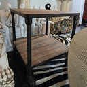 A rustic two-tier wooden end table with dark metal legs, styled in a room with classical statues and a zebra rug.
