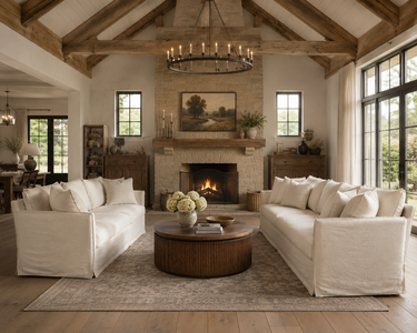 A rustic, high-ceilinged living room with exposed wood beams, a stone fireplace, and a round wood coffee table centered between two off-white slipcovered sofas on a large patterned rug.