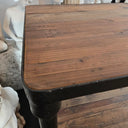 A rustic wooden table with a dark metal frame and leg, shown in a home setting with decorative white statues in the background.