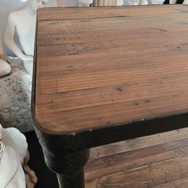 A rustic wooden table with a dark metal frame and leg, shown in a home setting with decorative white statues in the background.