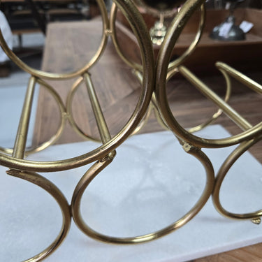 A close-up of a gold metal wine bottle rack with circular interlocking rings, resting on a white marble slab over a wooden floor in what appears to be a showroom or furniture store.