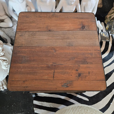 A square wooden end table with a rustic medium-brown finish and visible wood grain, photographed from above in an interior setting with white figurines, a zebra-pattern rug, and decor in the background.